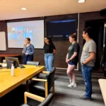 Outreach Four researchers standing in a boardroom during a hybrid lab meeting with Sollio, with remote participants projected on a large screen.