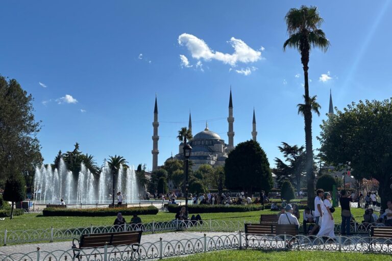 Sunny outdoor view of Sultanahmet Mosque (Blue Mosque) in Istanbul framed by tall minarets, palm trees, green lawns, and a fountain spraying water under a bright blue sky, with visitors walking and sitting on park benches.