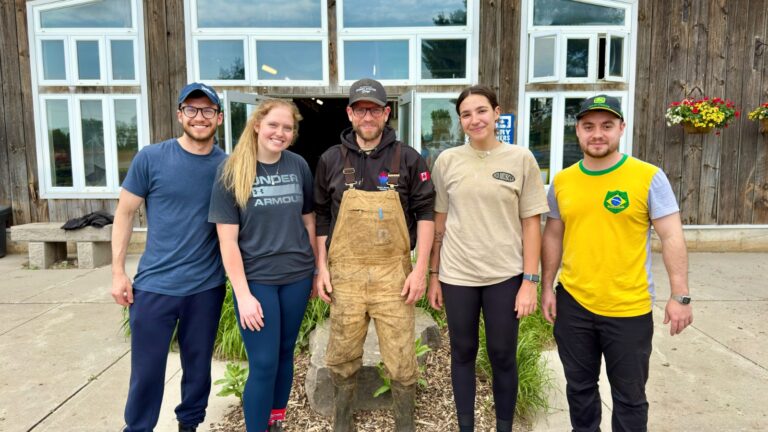 Group photo of five researchers standing together in front of a wooden farm building with large glass windows.