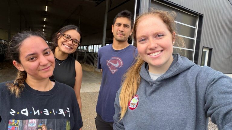 Selfie of four young researchers smiling outside a large farm building, with a red-haired woman in a grey hoodie in the foreground.